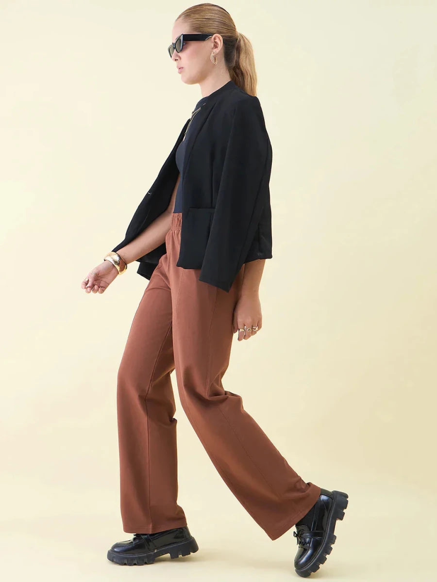 Woman in sunglasses, black blazer, and 98degreenorth Dark Rust Wide Leg Pintuck Trackpants walks by a light background.