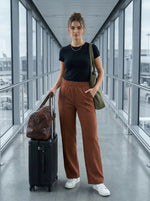 Woman in an airport, with luggage, wears black shirt and 98degreenorth Dark Rust Wide Leg Pintuck Trackpants.