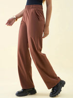 Model in 98degreenorth Dark Rust Wide Leg Pintuck Trackpants, black top, chunky shoes & gold bracelet on light backdrop.