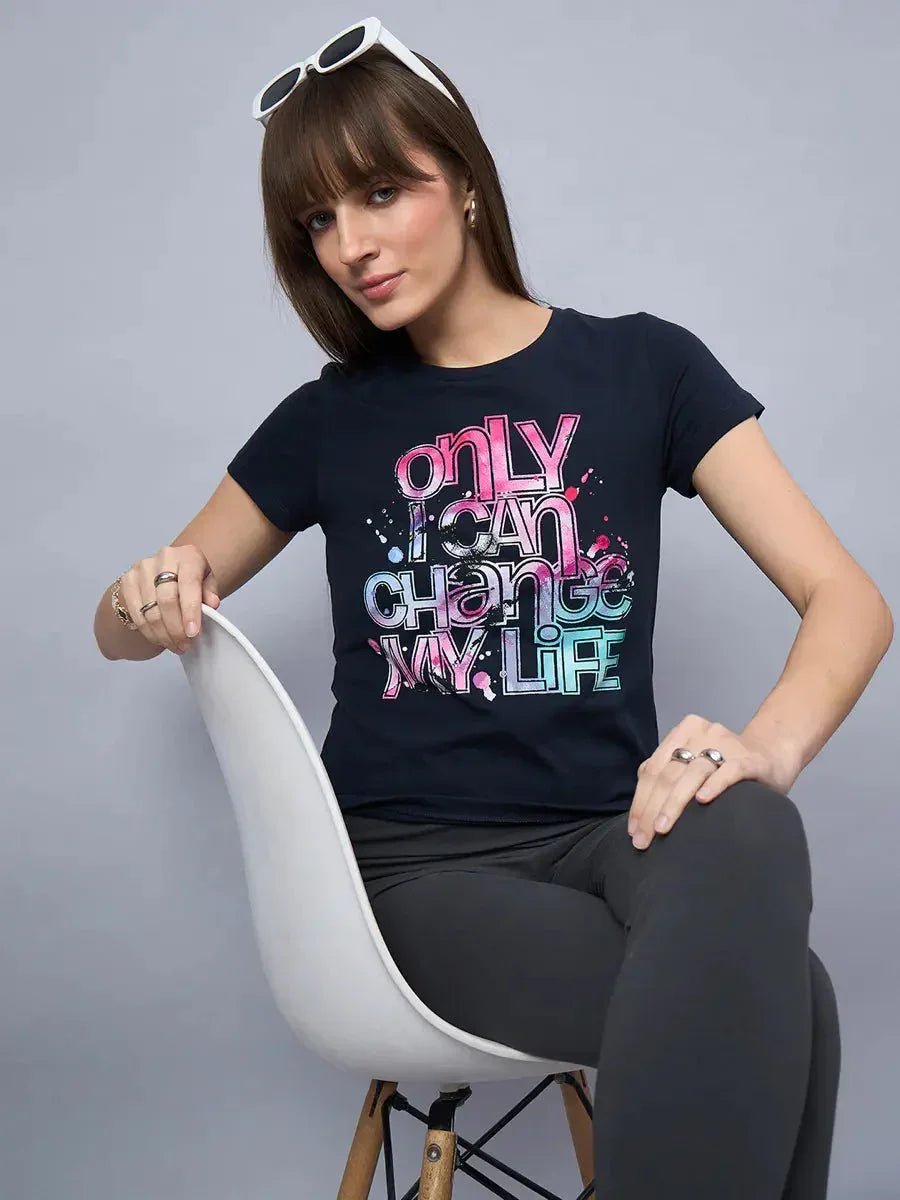 Woman in a 98degreenorth soft cotton typography t-shirt, seated on a white chair against a gray background.