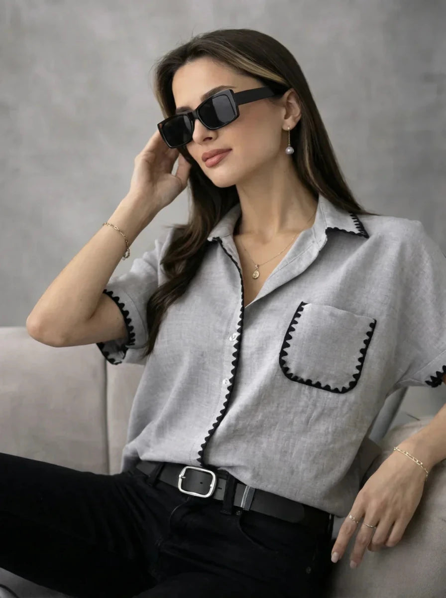 Woman wears 98degreenorth’s Grey Whipstitch Trim Cotton Shirt, black sunglasses, and pants on a light sofa.