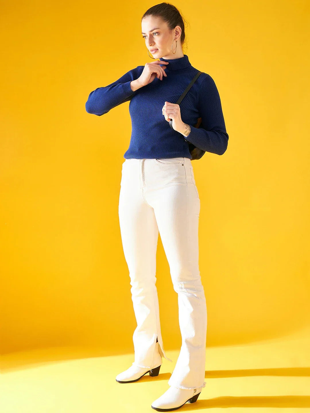 Woman in a 98degreenorth Indigo Ribbed Knit Turtleneck Sweater, white pants, and black backpack against yellow background.