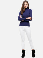 Woman in a 98degreenorth Ink Blue Ribbed Turtleneck Sweater, white pants, and black boots stands smiling with arms crossed.