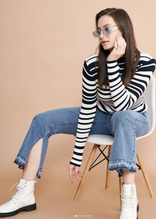Woman in striped sweater, blue jeans, and white boots sits on chair, wearing blue sunglasses, against beige background.