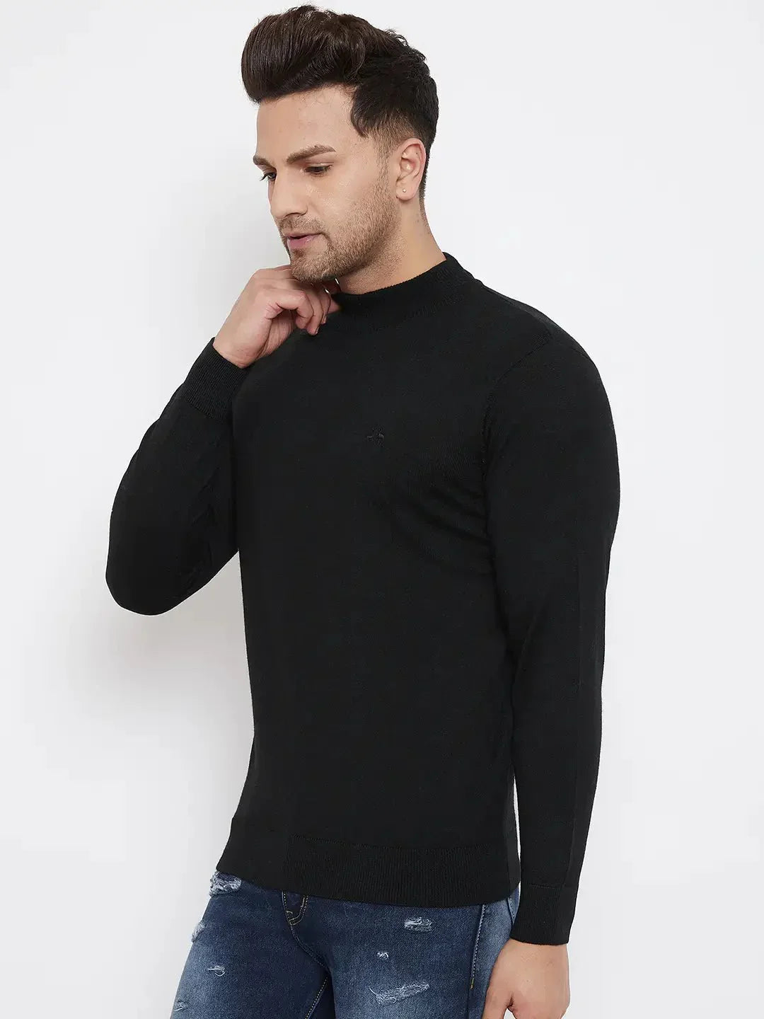 A man in a 98degreenorth Men Merino Wool Black Turtleneck Pullover and blue distressed jeans stands against a plain white background, looking down with one hand on his chin.