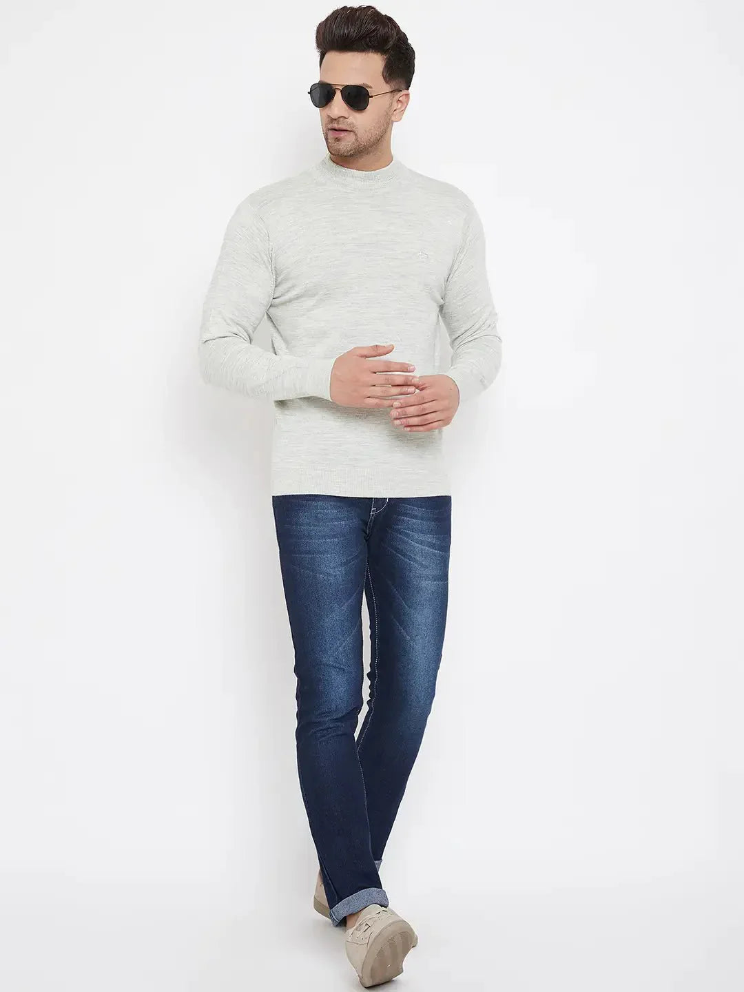 A man in dark sunglasses wears the 98degreenorth Men Merino Wool Ecru Turtleneck Pullover with blue jeans and beige shoes, posing against a plain white background with relaxed arms and one leg crossed in front of the other.