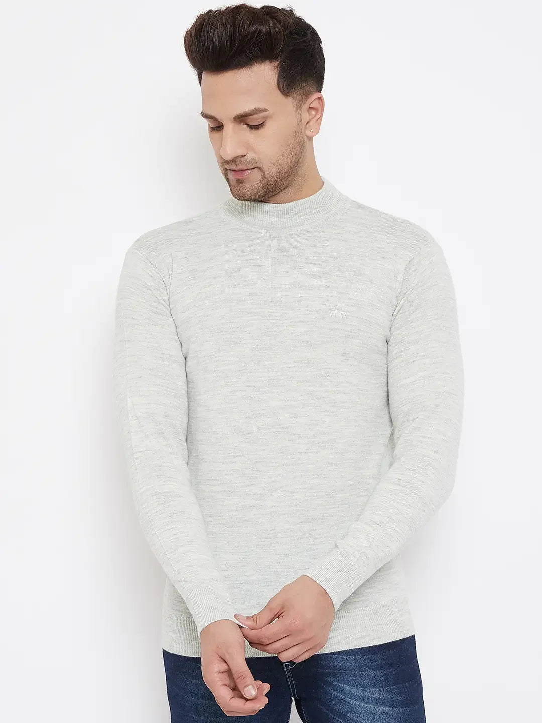 A man with short dark hair wears the 98degreenorth Men Merino Wool Ecru Turtleneck Pullover and blue jeans, standing against a plain white background with his hands clasped, looking down.