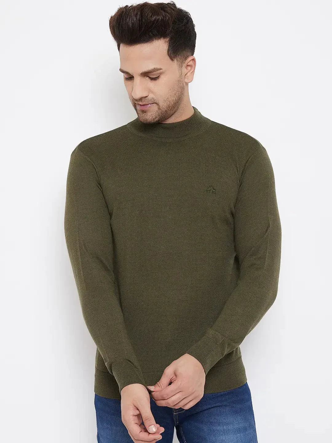 A man wearing the 98degreenorth Men Merino Wool Olive Turtleneck Pullover with blue jeans stands against a white background, looking down with his hands clasped in front of him.