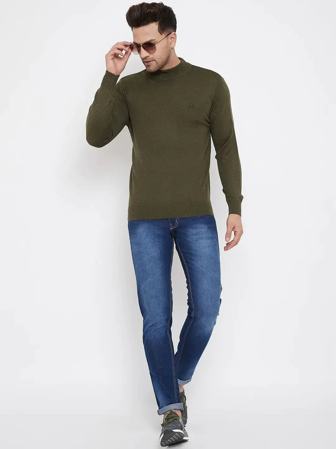A man wearing sunglasses, blue jeans, gray sneakers, and a 98degreenorth Men Merino Wool Olive Turtleneck Pullover walks confidently against a plain white background.