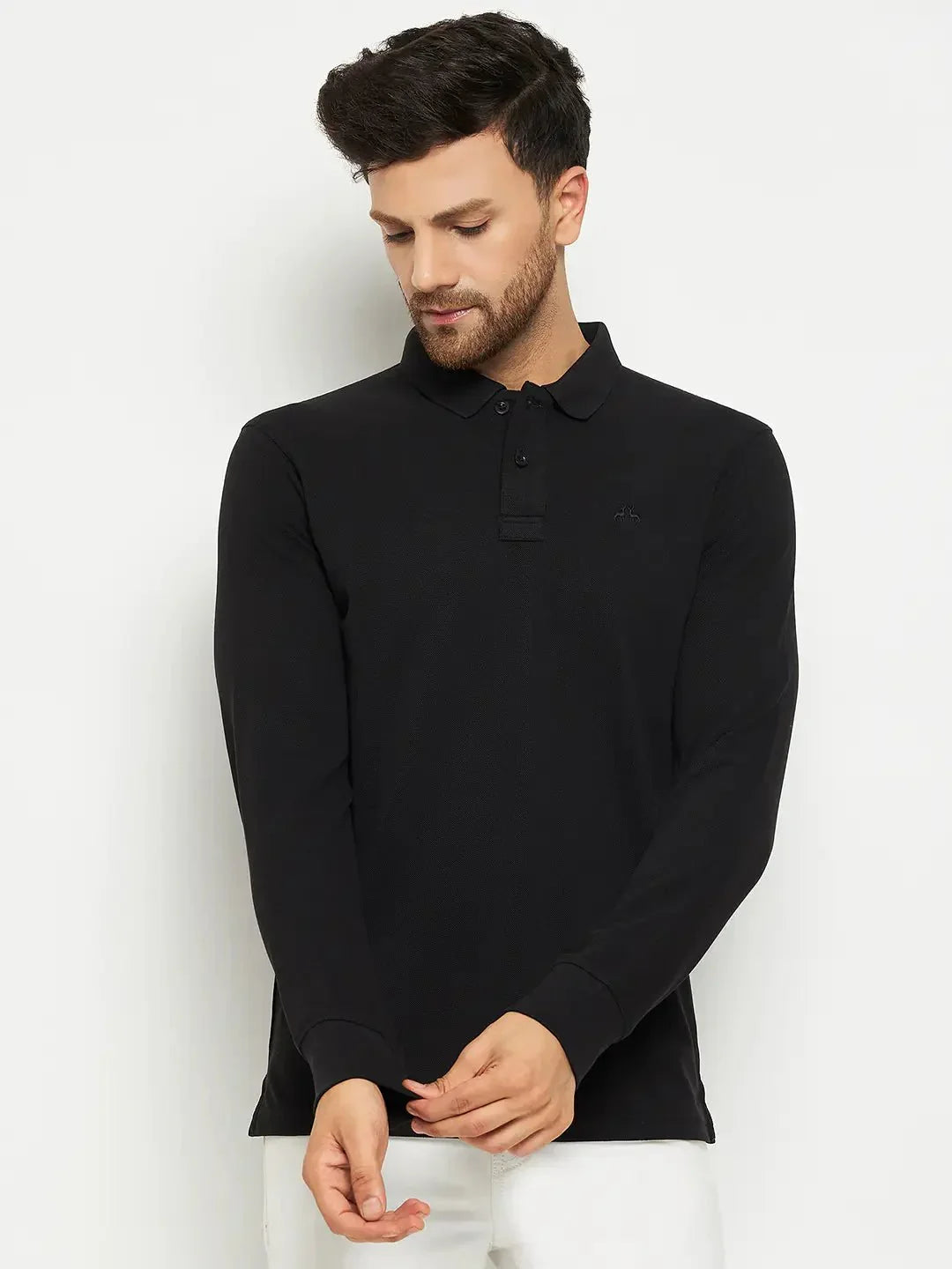 Man in a 98degreenorth Men Polo Collar Full Sleeves Black T-shirt and white pants adjusts his sleeve against a plain backdrop.