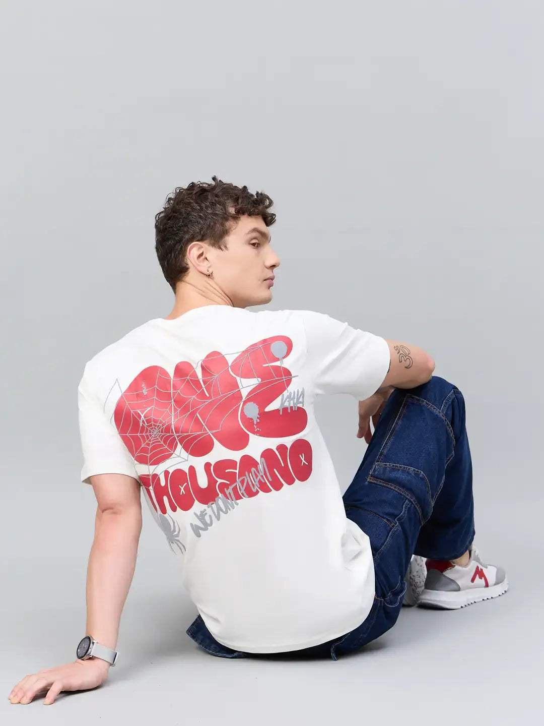 A person in a 98degreenorth Men Typography Offwhite Printed Round Neck T-shirt and jeans sits on the floor, glancing back.