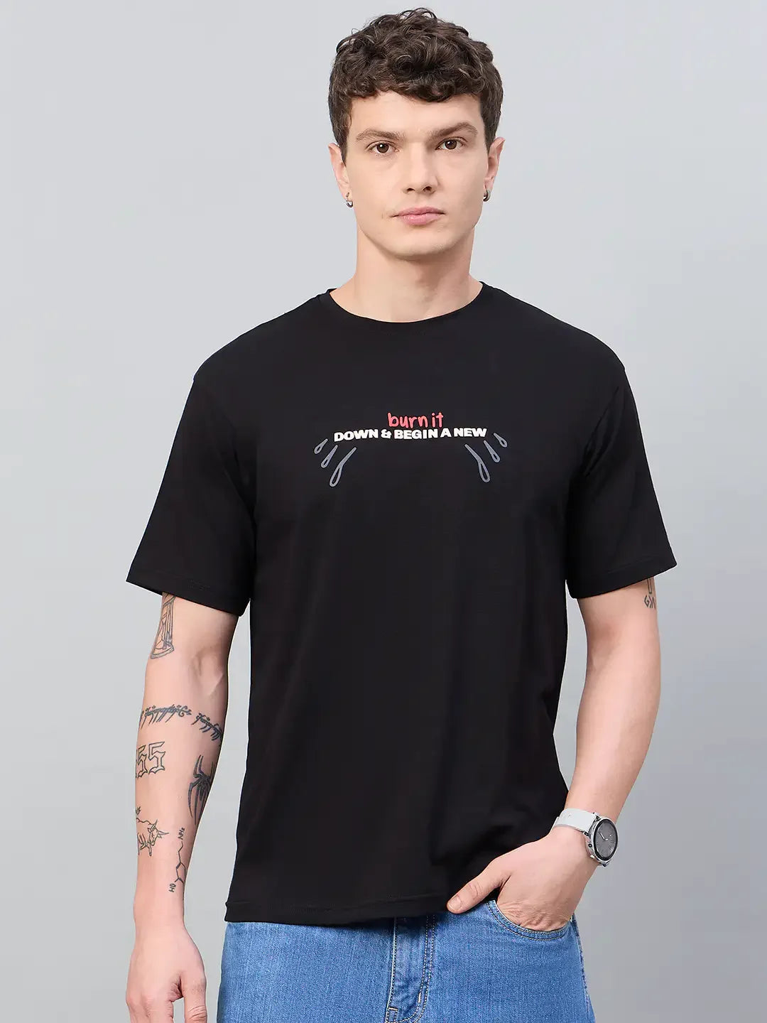 Man in a 98degreenorth Men Typography Printed Black Round Neck T-shirt, blue jeans, tattoos, and a watch.