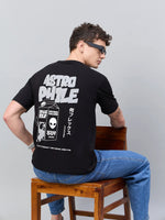 Man in sunglasses and jeans sits, revealing the 98degreenorth Men Typography Printed Round Neck Black T-shirt.