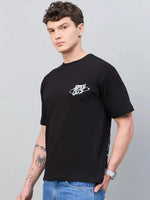 Man in a 98degreenorth Men Typography Printed Round Neck Black T-shirt and blue jeans stands against a gray background.