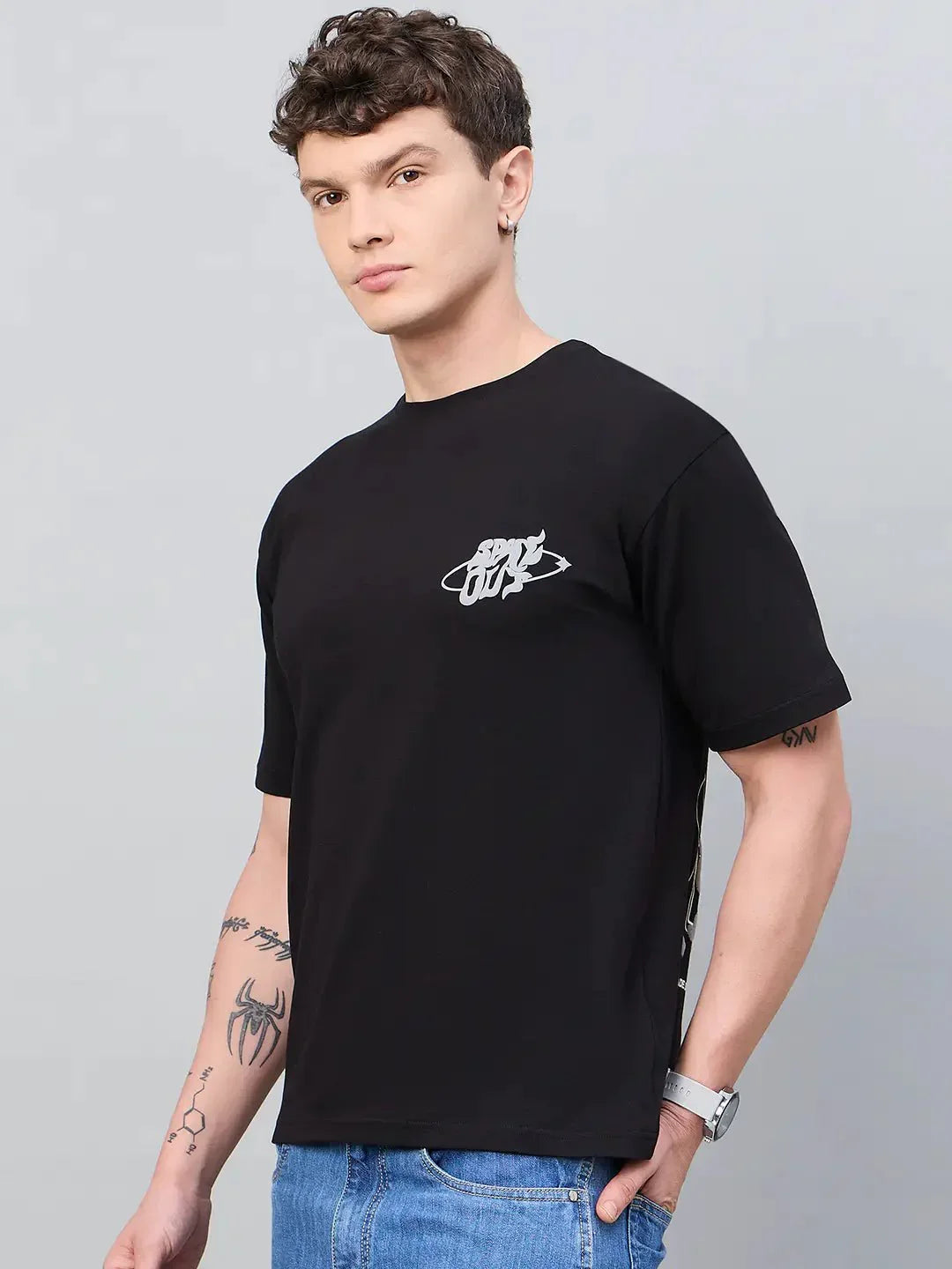 Man in a 98degreenorth Men Typography Printed Round Neck Black T-shirt and blue jeans stands against a gray background.