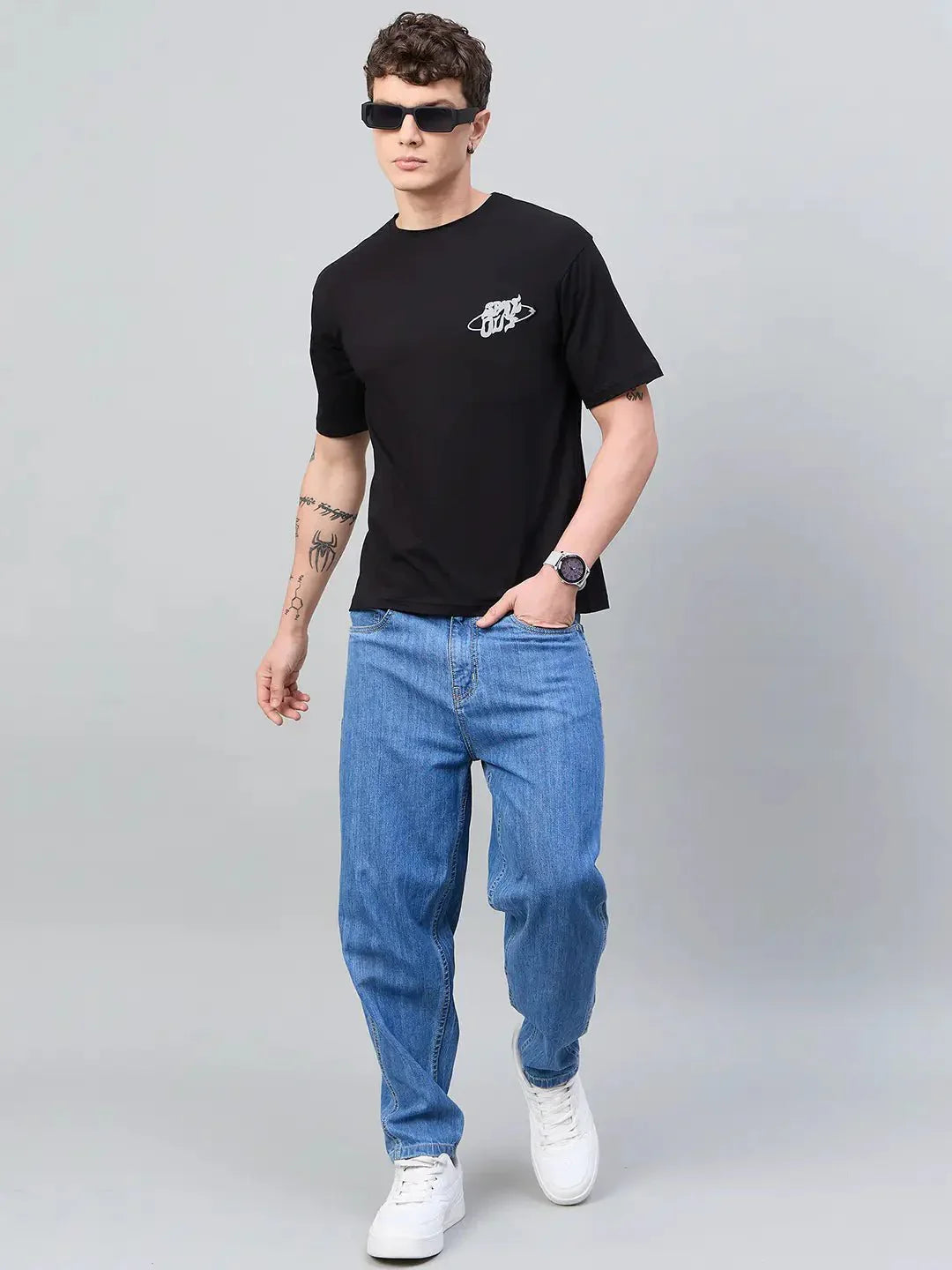 Man in 98degreenorth Men Typography Printed Round Neck Black T-shirt, blue jeans, white sneakers, sunglasses, gray backdrop.