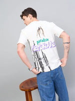 A man in jeans wears the 98degreenorth Men Typography Printed Round Neck Offwhite T-shirt, standing by a wooden stool.