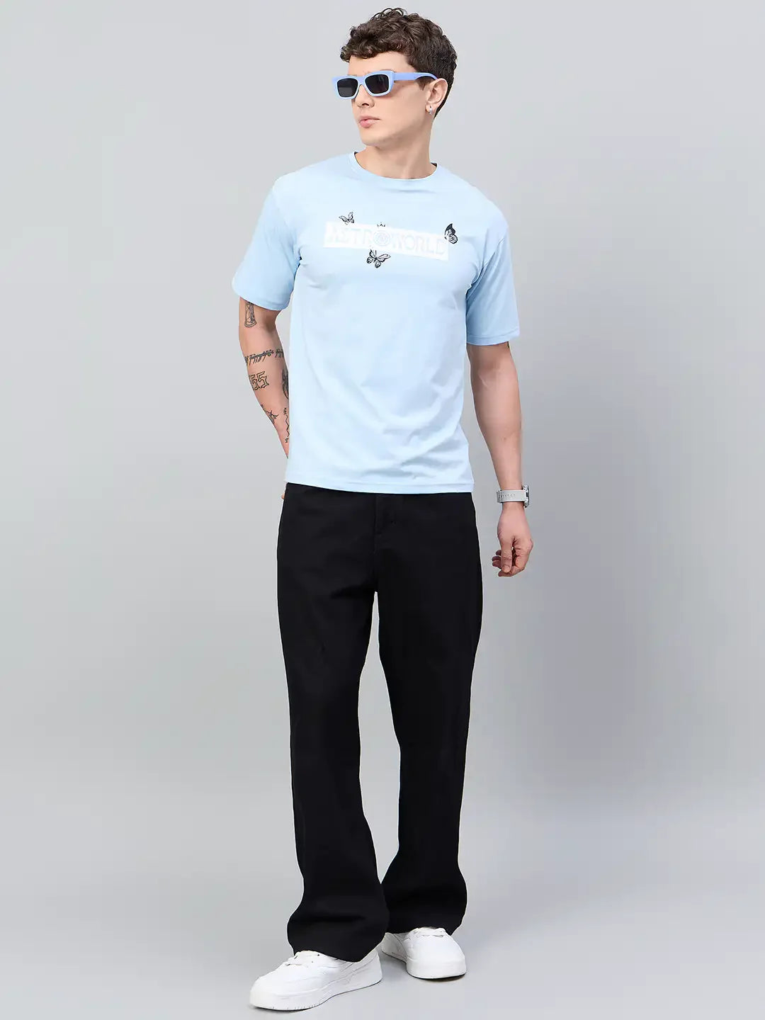 Man in 98degreenorth Men Typography Printed Round Neck Sky T-shirt, black pants, white sneakers, blue sunglasses.