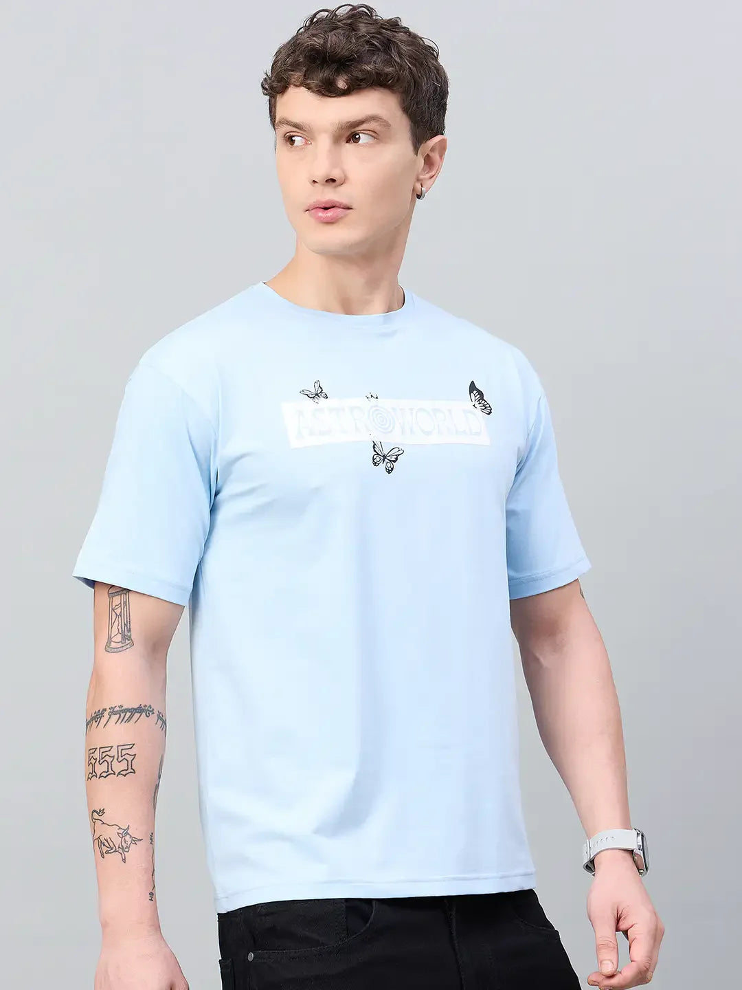 Man wears an oversized 98degreenorth Men Typography Printed Round Neck Sky T-shirt, streetwear style, on gray background.