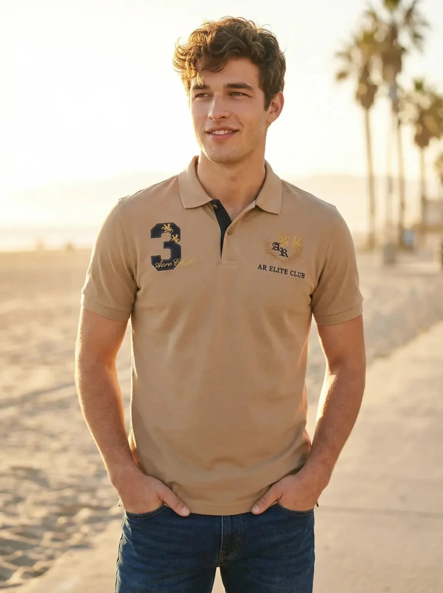 Man in a 98degreenorth Men's Beige Aero Club No.3 Polo T-Shirt and jeans stands on a beach path with palm trees.