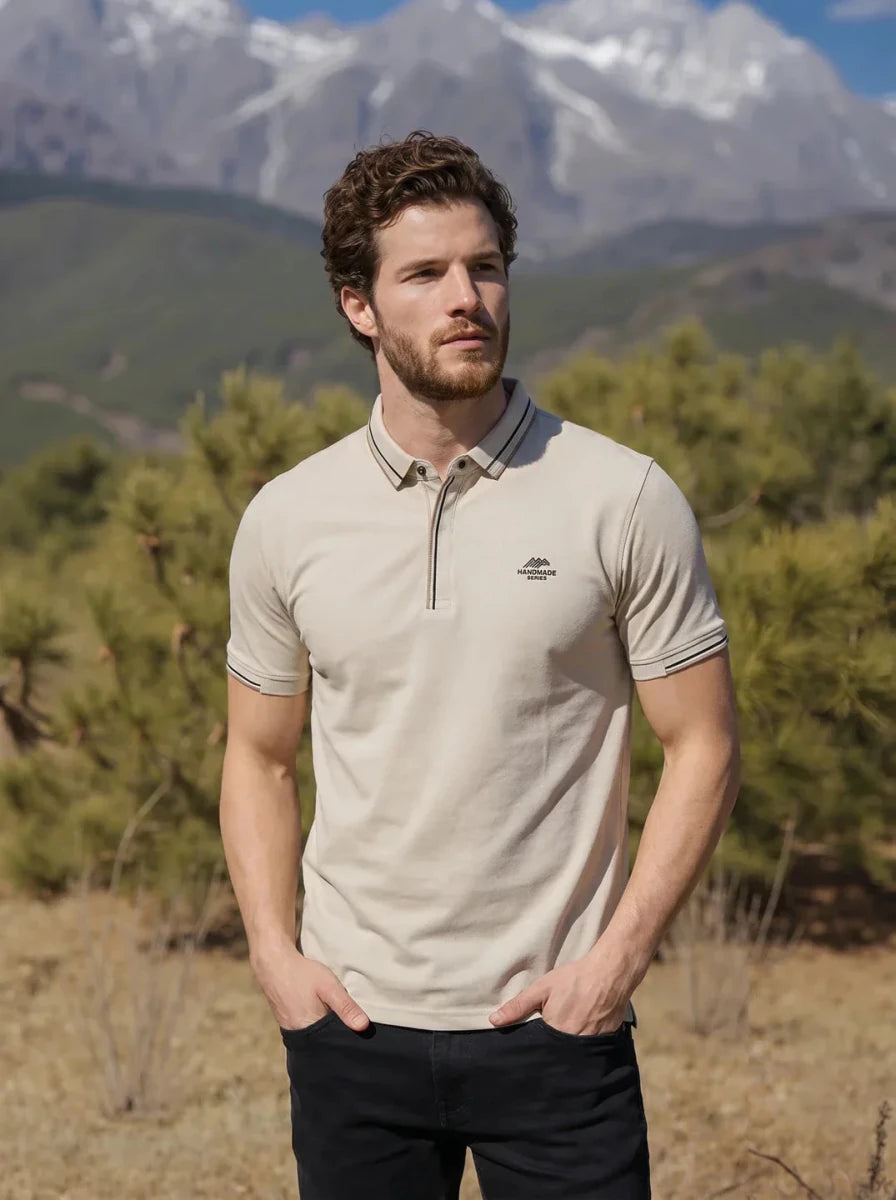 A man in a 98degreenorth Men's Beige Polo T-Shirt stands outdoors with trees and mountains behind him.