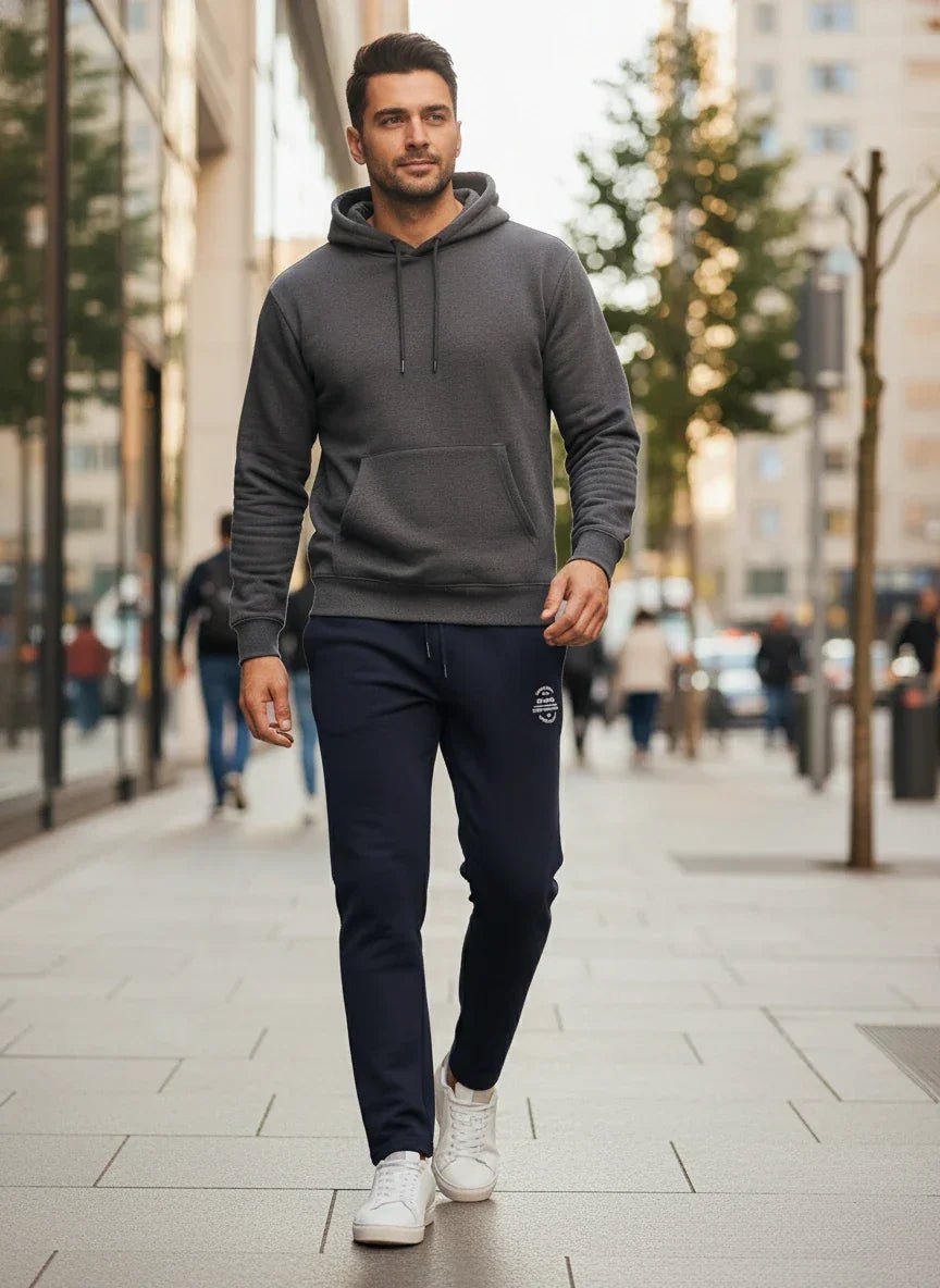 A man in 98degreenorth Men’s Black Fleece Trackpants walks on a city sidewalk with buildings and trees in view.