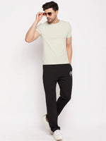 Man wears 98degreenorth Men's Black French Terry Lycra Trackpants, sunglasses, beige tee & white sneakers against plain backdrop.
