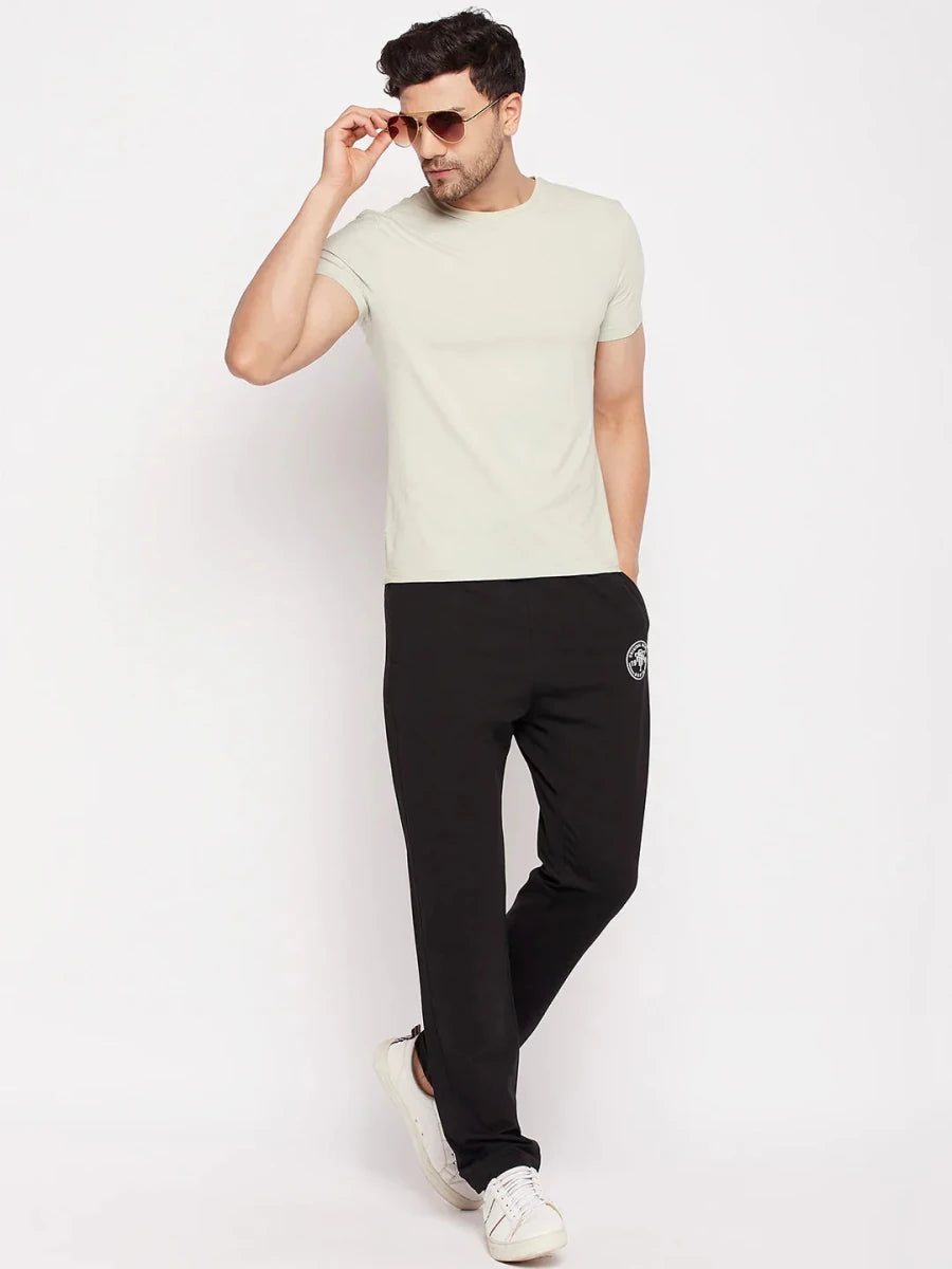 Man wears 98degreenorth Men's Black French Terry Lycra Trackpants, sunglasses, beige tee & white sneakers against plain backdrop.
