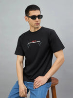Man in sunglasses wearing the 98degreenorth Men's Black Graphic Oversized T-Shirt, sitting on a stool against gray.