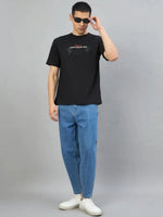 Man in sunglasses wears a 98degreenorth Black Graphic Oversized T-Shirt, blue jeans, and white sneakers against gray background.
