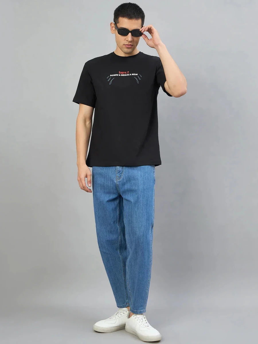 Man in sunglasses wears a 98degreenorth Black Graphic Oversized T-Shirt, blue jeans, and white sneakers against gray background.