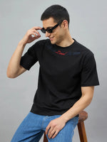 Man in a 98degreenorth black graphic oversized t-shirt with gothic cross print, sitting and adjusting his sunglasses.