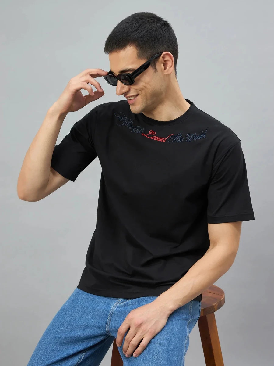Man in a 98degreenorth black graphic oversized t-shirt with gothic cross print, sitting and adjusting his sunglasses.