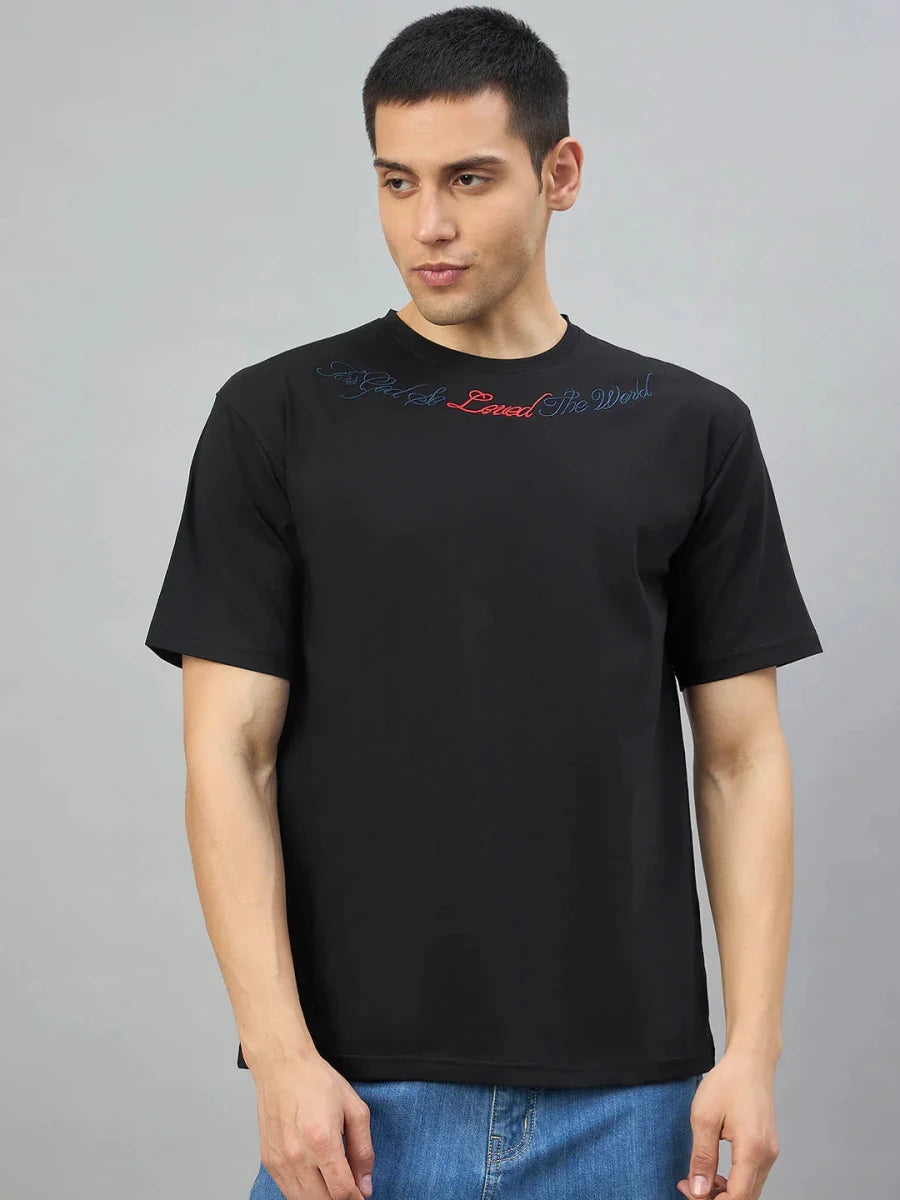 Man in a 98degreenorth black oversized T-shirt with Gothic Cross embroidery, against a plain gray background.