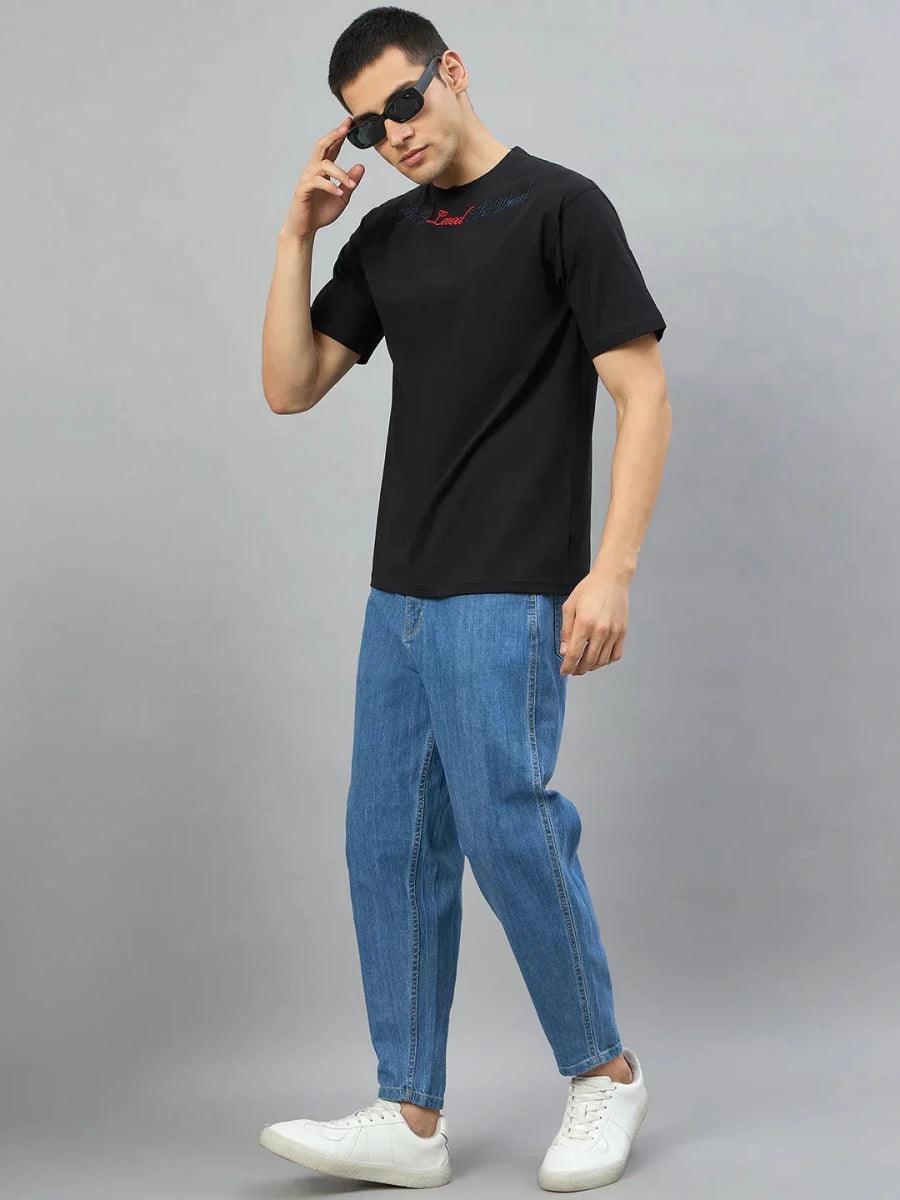 Man in 98degreenorth Men's Black Graphic Oversized T-Shirt with Gothic Cross Embroidery, posing against gray background.