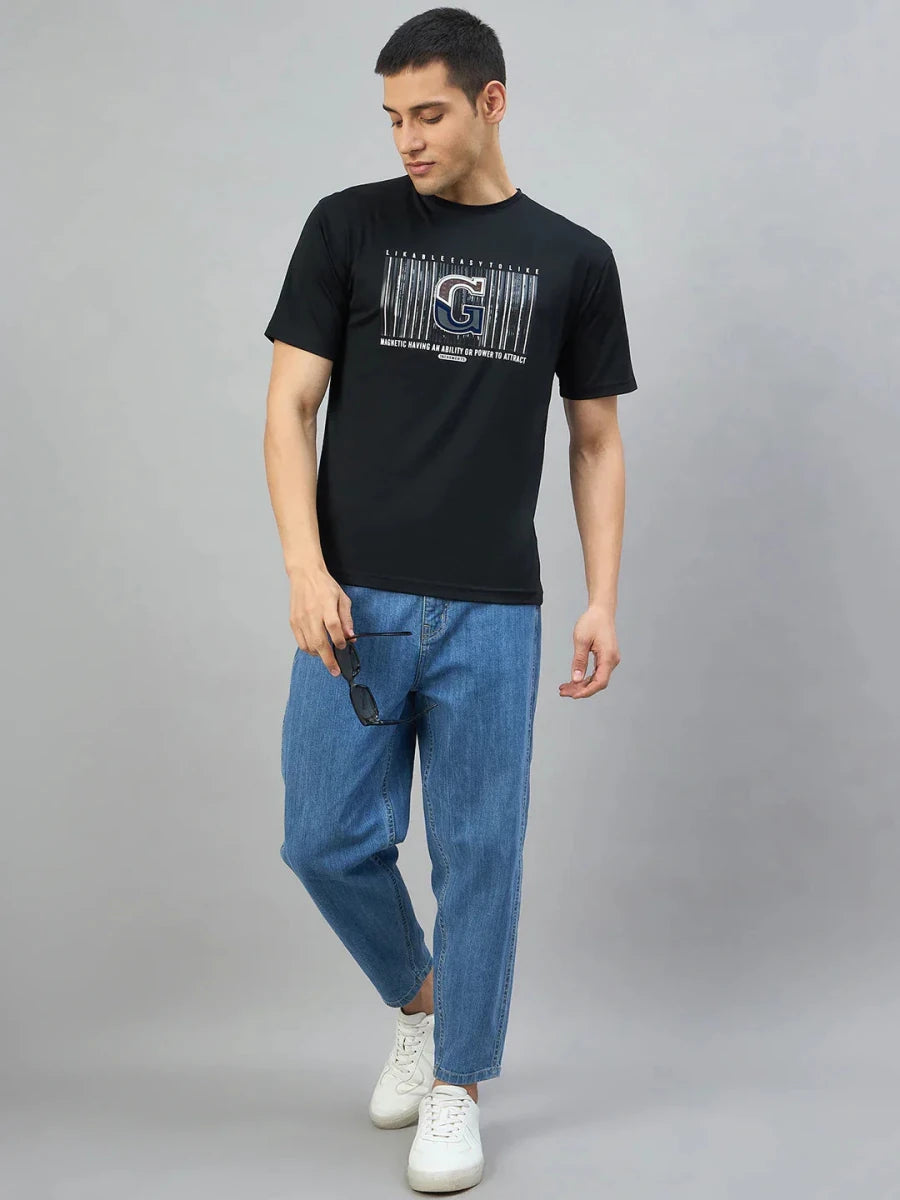 Man in 98degreenorth Men's Black Magnetic Graphic T-Shirt, blue jeans, and sneakers holds sunglasses against gray background.