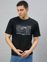 Man wears 98degreenorth Men's Black Magnetic Graphic T-Shirt, standing against a grey background, looking down.