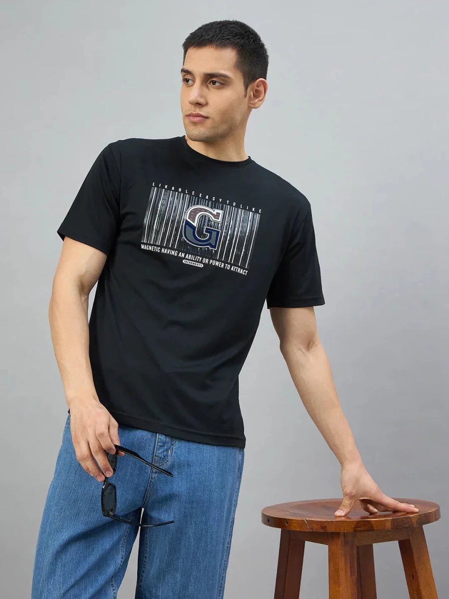 Man wears a 98degreenorth Men's Black Magnetic Graphic T-Shirt with blue jeans, standing by a wooden stool holding sunglasses.