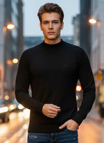 A man wears a 98degreenorth Men’s Black Merino Wool Turtle Neck Sweater on a city street at dusk with blurred lights behind.