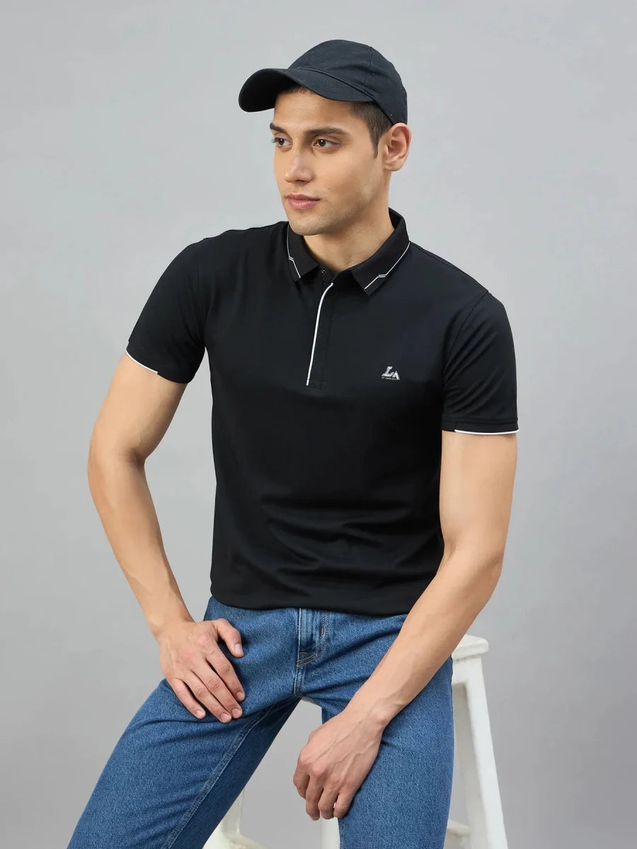 Man in a 98degreenorth black polo with white tipping, blue jeans, and a black cap sits on a white stool against gray.