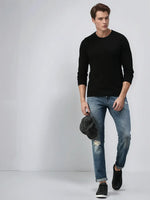 Man in a 98degreenorth Black Premium Merino Wool Sweater, blue ripped jeans, and black shoes holds a cap.