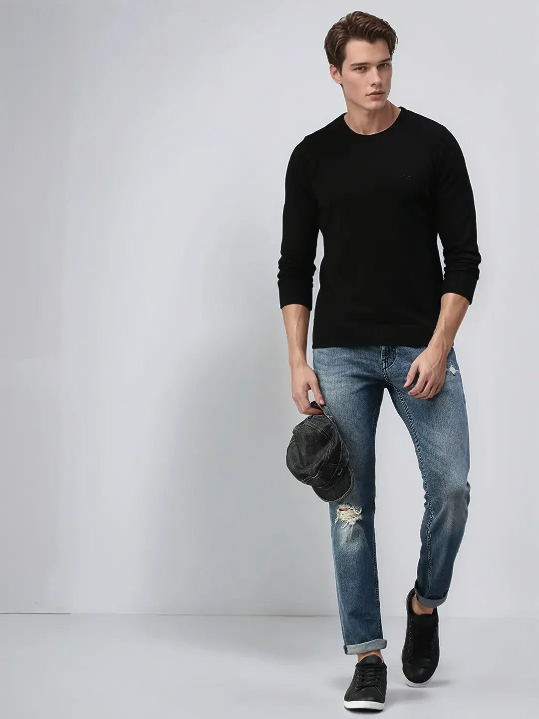 Man in a 98degreenorth Black Premium Merino Wool Sweater, blue ripped jeans, and black shoes holds a cap.