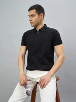 A man wears the 98degreenorth Men's Black Quarter Zip Polo T-Shirt, sitting on a stool and holding sunglasses.