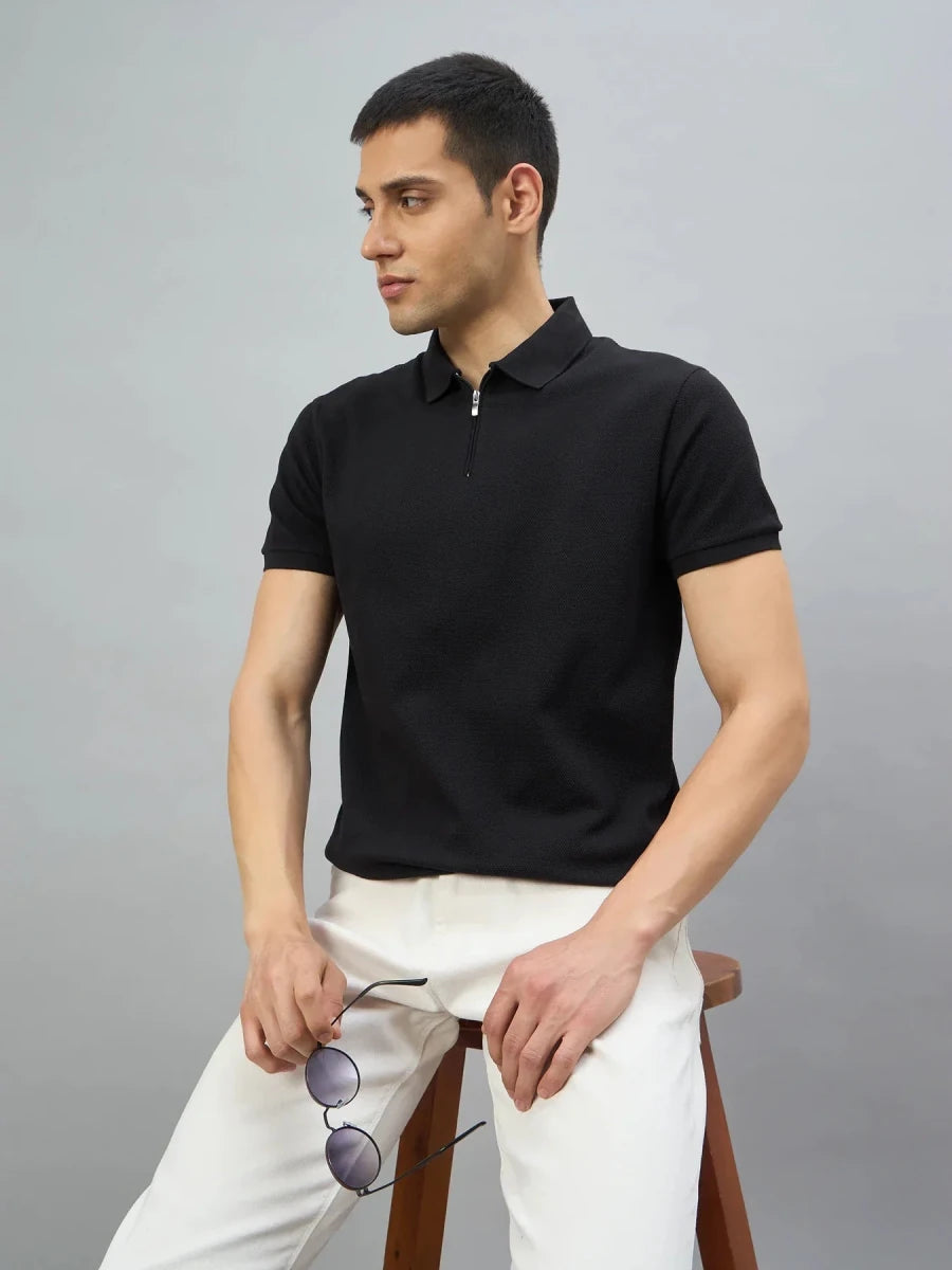 A man wears the 98degreenorth Men's Black Quarter Zip Polo T-Shirt, sitting on a stool and holding sunglasses.