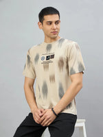 A man in a 98degreenorth Men's Camel Abstract Ikat Print T-Shirt and black pants sits on a stool, looking left.