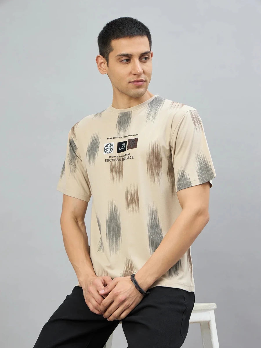 A man in a 98degreenorth Men's Camel Abstract Ikat Print T-Shirt and black pants sits on a stool, looking left.