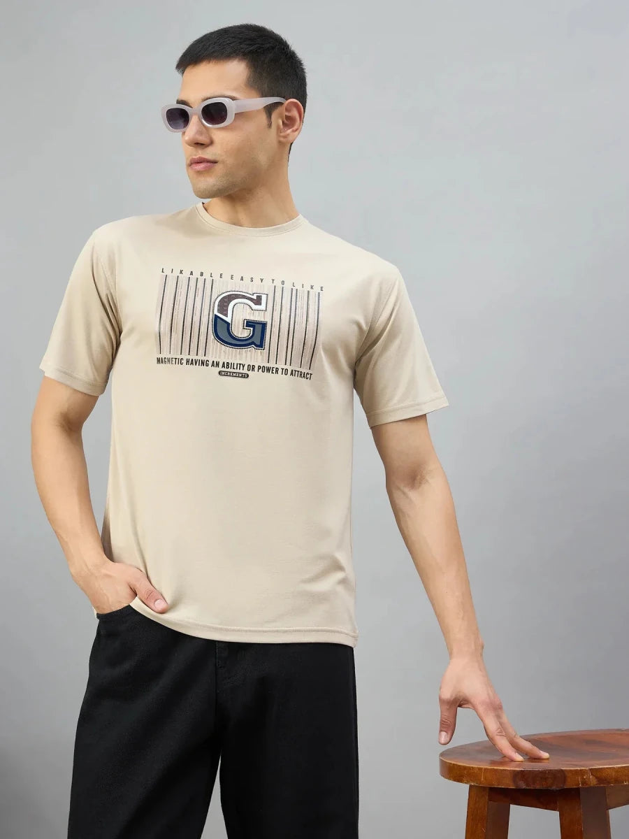 Man in 98degreenorth camel varsity G graphic oversized t-shirt stands with sunglasses, hand in pocket, by a wooden stool.