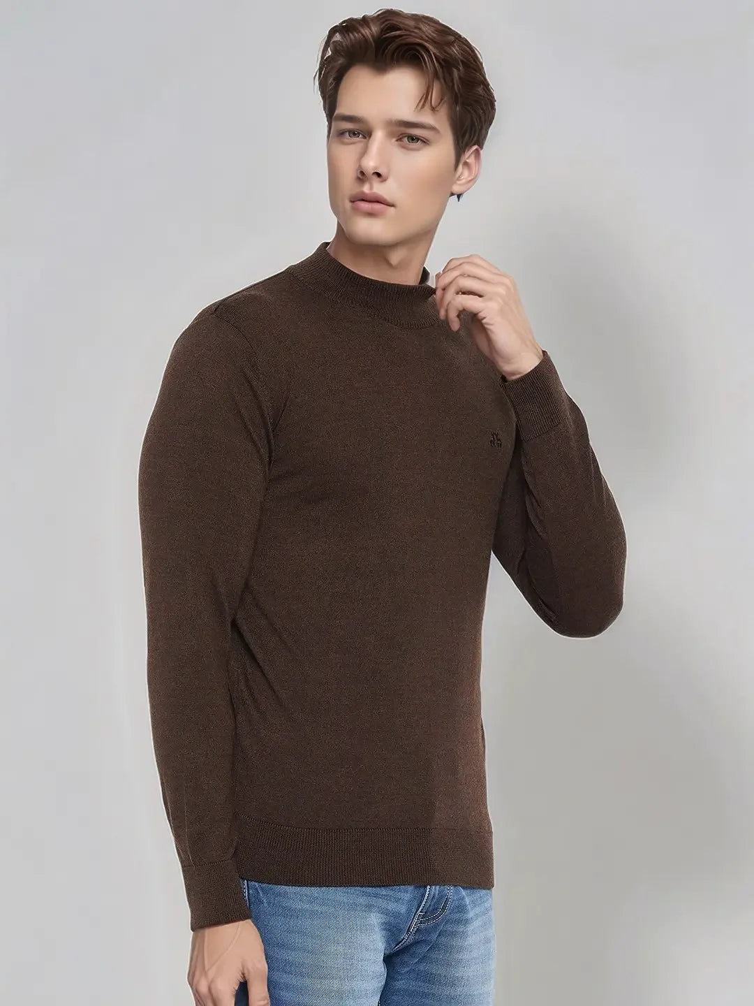A man models the 98degreenorth Men’s Coffee Merino Wool Turtle Neck Sweater with blue jeans on a light gray background.