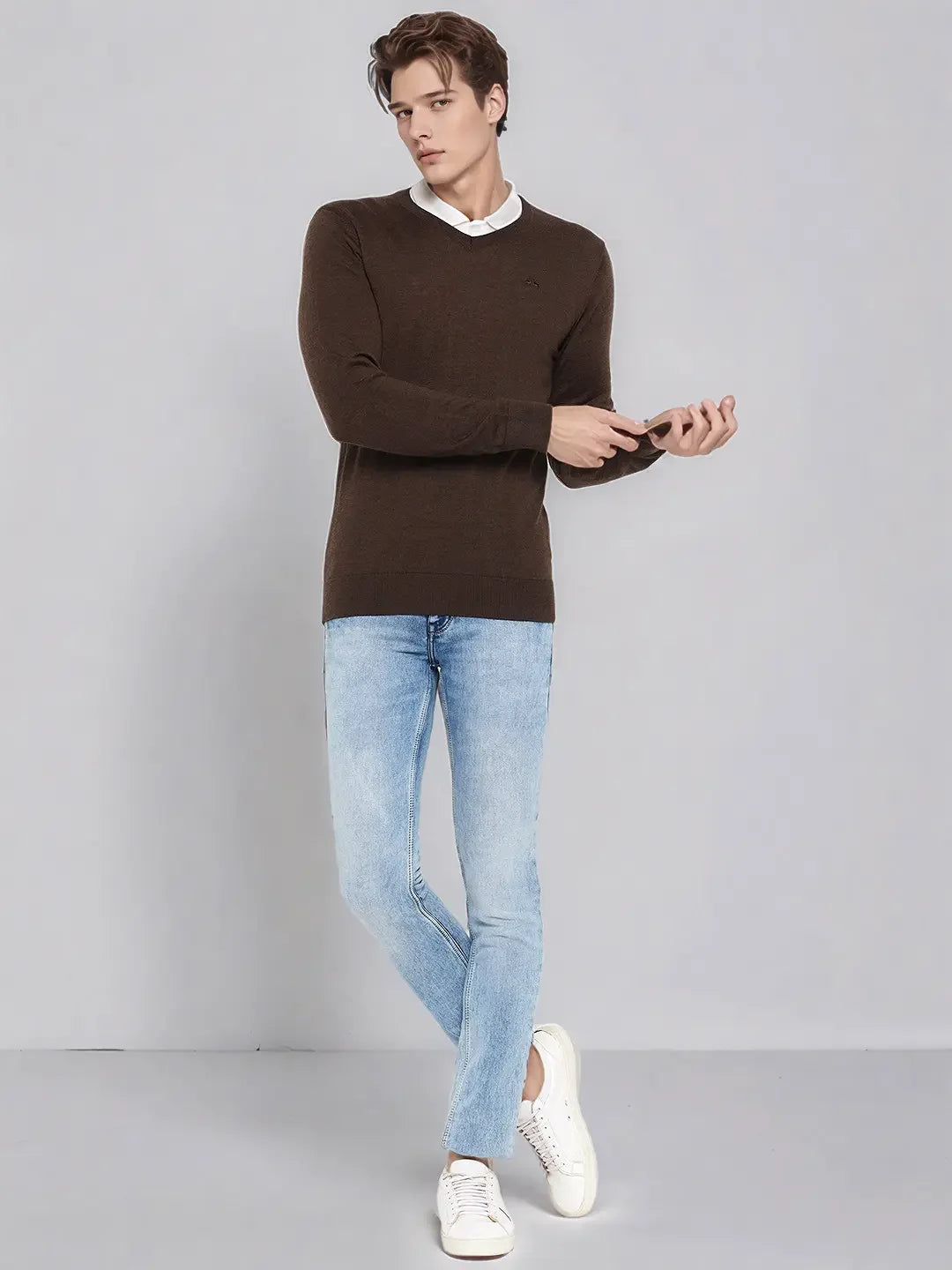 Man wears 98degreenorth Men’s Coffee Merino Wool V-Neck Sweater, light blue jeans, and white sneakers on a gray backdrop.
