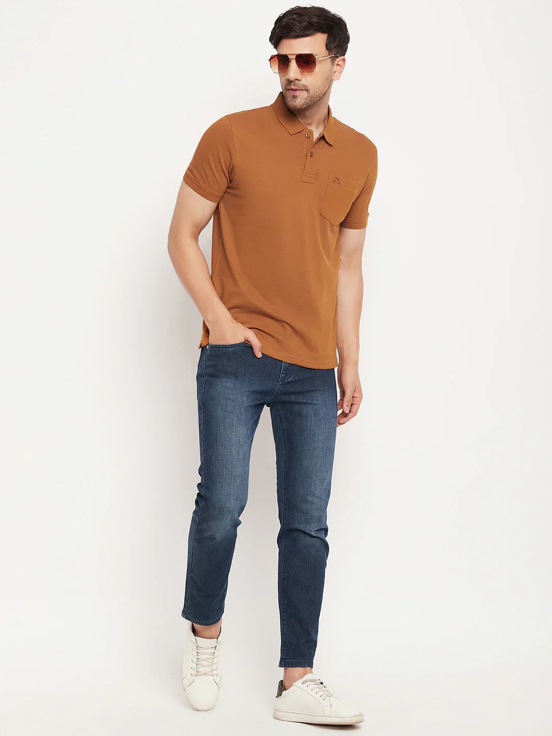 Man wears 98degreenorth Men's Copper Polo T-Shirt, blue jeans, white sneakers & sunglasses against white background.